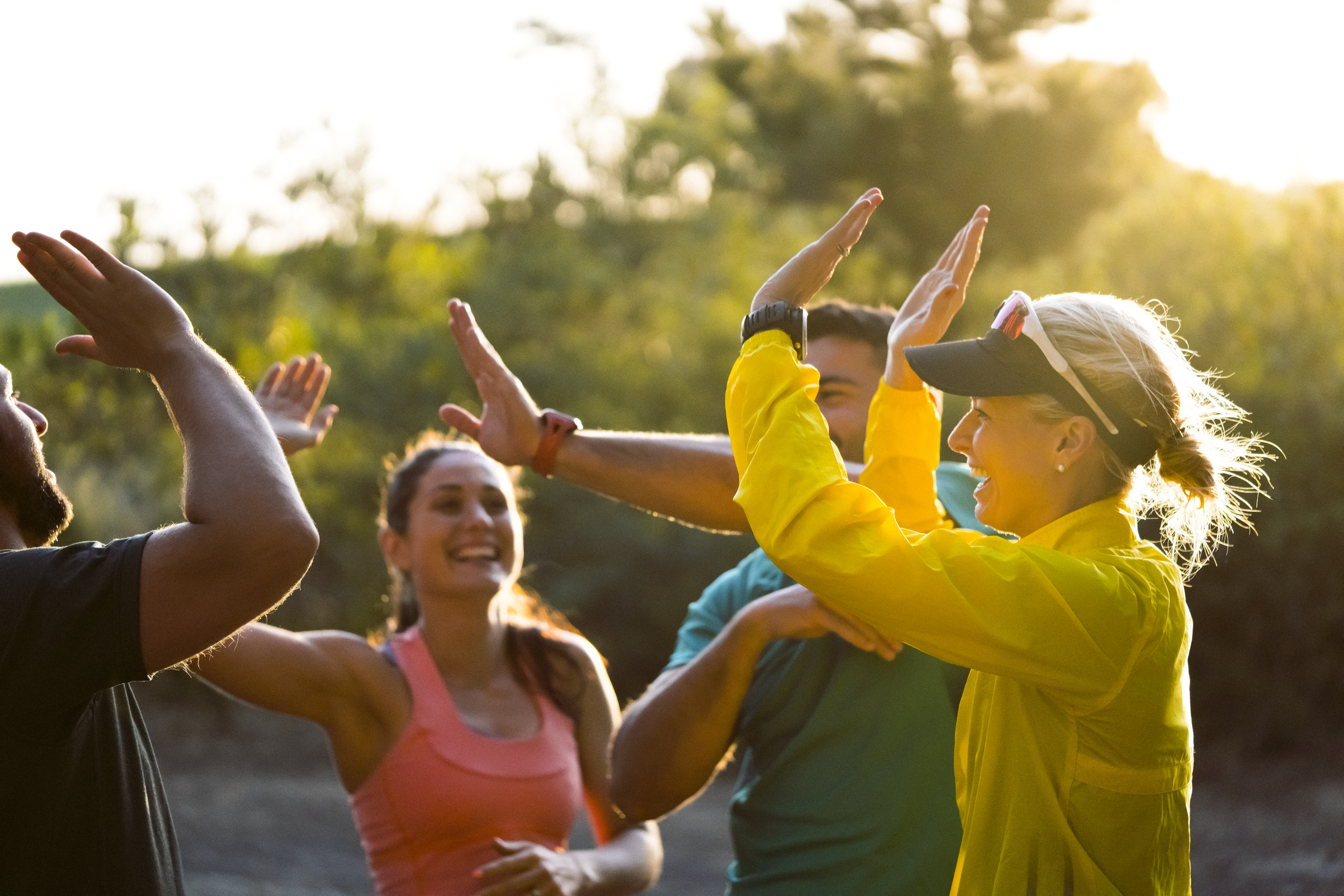 A photo of cheerful friends giving high-five. Happy men and women are wearing sportswear. They are in park on sunny day.