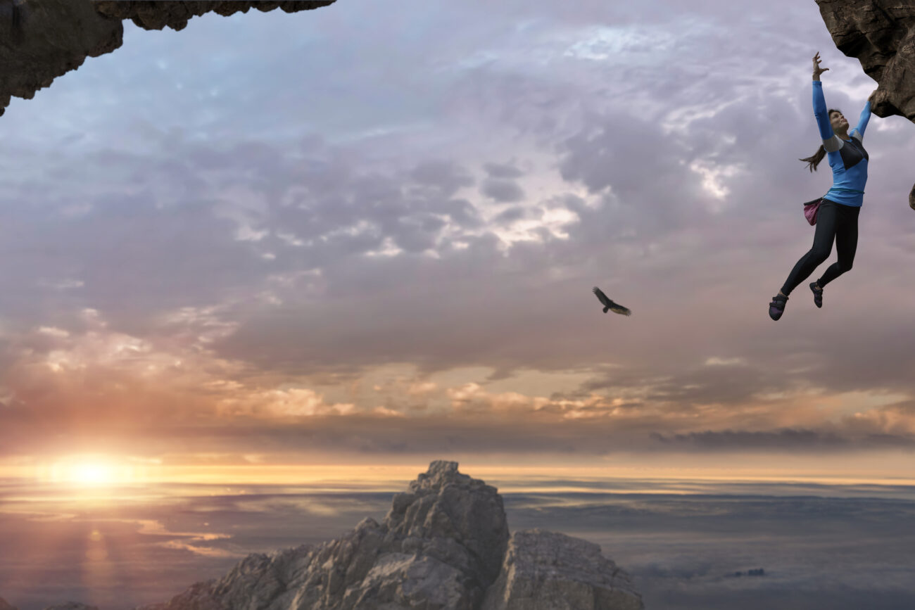 A young woman wearing climbing leggings, top, climbing shoes with chalk bag hanging from a rock face with one arm and reaching for a hold with the other. The rock climber is ascending a sheer rock face, climbing high up, close to other mountain peaks under a beautiful dawn sky.